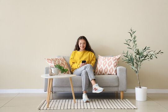 Young Asian woman using mobile phone on grey sofa near beige wall