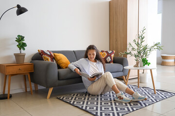 Young Asian woman reading magazine on carpet near black sofa at home