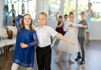 Smiling junior schoolchildren in party dresses learning dancing waltz with pedagogue in social hall