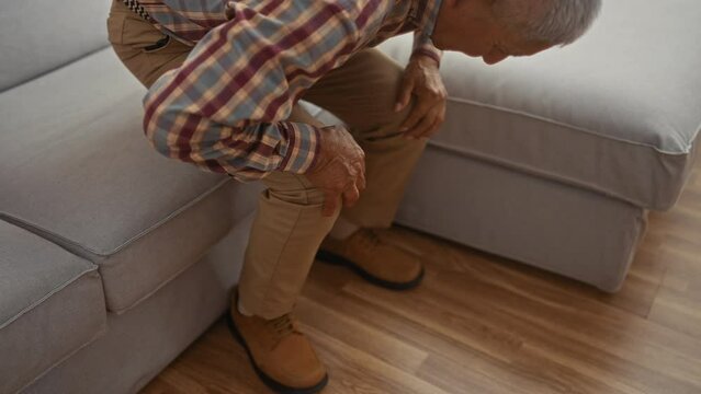 Mature hispanic man with grey hair and plaid shirt clutching his knees while standing up from a sofa in a cozy living room.