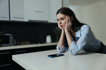 Stressed woman sitting at kitchen counter with head in hands and cell phone in front of her,...