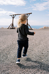Little girl goes to a wooden swing on a seashore. Back view