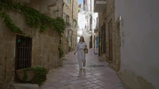 A young hispanic woman explores the charming streets of polignano a mare in puglia, italy, surrounded by historic stone buildings and vibrant greenery.