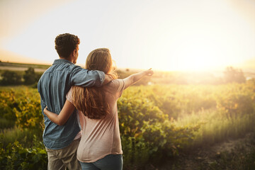 Back, hug and couple in sunrise, pointing and together with care, love and date for anniversary in outdoor. Morning, partner and person with embrace, nature and peace in field and relax in Texas