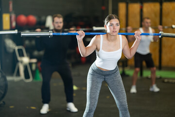 Fototapeta premium Focused motivated sporty young girl performing set of exercises with barbell during intense group workout. Concept of weight training ....