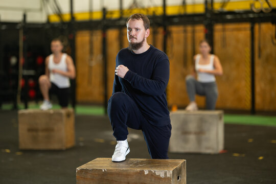 Active young bearded man performing step-ups on wooden plyo box during group fitness workout, concentrating on working lower body muscles and improving core stability