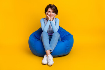 Fototapeta premium Full size photo of adorable peaceful girl sit comfort bag hands touch cheeks isolated on yellow color background
