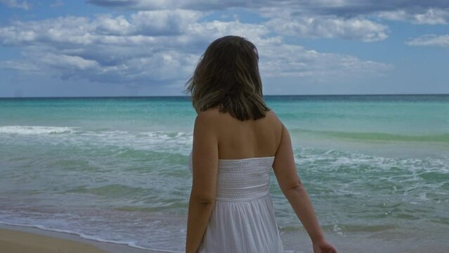 A young hispanic woman in a white dress enjoying the beach at pescoluse in puglia, italy, with turquoise water and a cloudy sky.