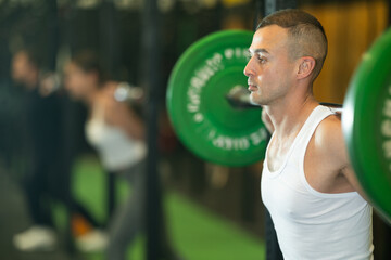 Athletic young adult man participating in intense group training in gym, performing exercises with...