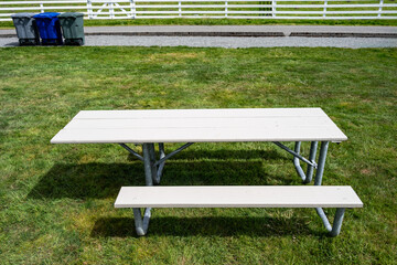 Setting up for a spring celebration in a grass field in the park, wheelchair accessible picnic table with garbage and recycle cans by white fence in background, sunny day outside
