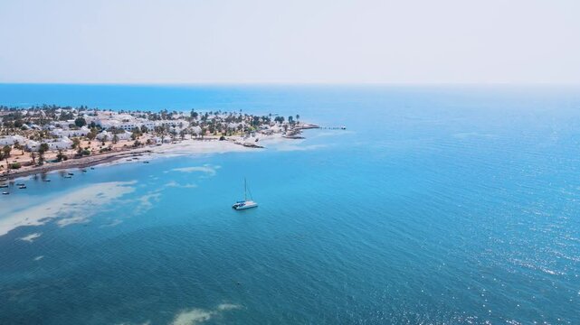 An aerial view of a sailboat is floating on a large body of water, surrounded by a beautiful beach with palm trees and houses nearby. at Tunisia Djerba