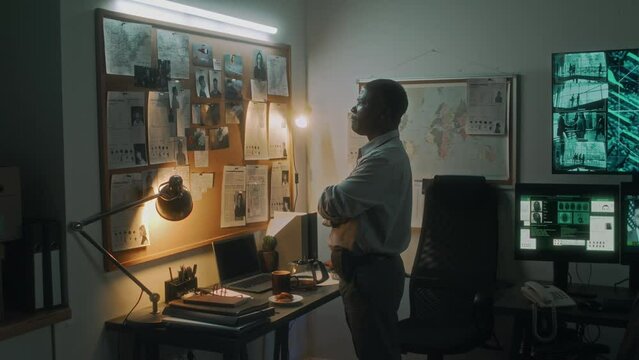 Medium full shot of African American federal agent standing in office in front of wall board with photos of murder scene and possible suspects, examining clues, while investigating homicide case