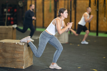 Concentrated young girl in fitness attire performing Bulgarian split squats using wooden plyometric box at gym. Bodyweight training concept