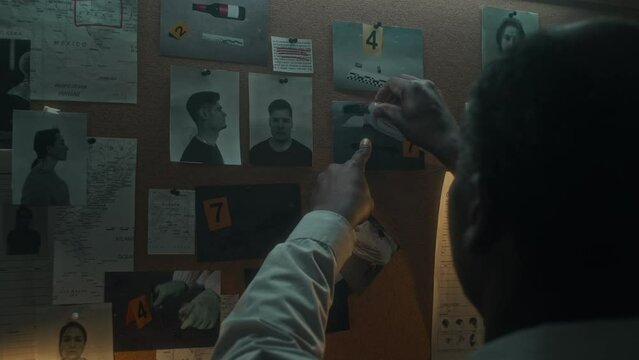 Close-up over-shoulder shot of black male agent pinning crime scene photos of victims body, forensic expert collecting evidence, onto wall board in office, next to pictures of convicted offenders