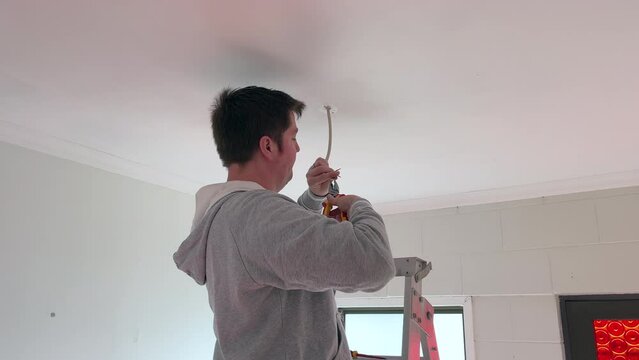 Middle-aged caucasian man using electrical pliers to strip and twist the insulation from copper wires hanging down from a lighting outlet in the ceiling of a 1970's house in New Zealand