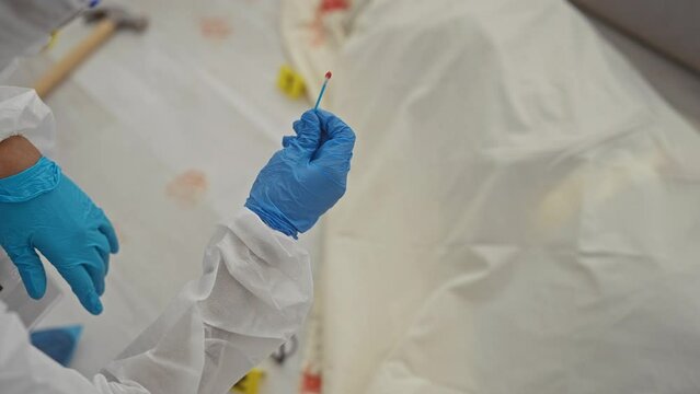 A forensic analyst in protective gear holds a swab at a crime scene with bloodstains.