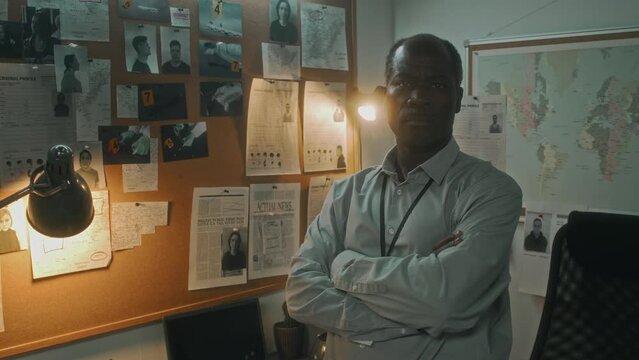 Medium portrait of black male police detective or special agent with folded arms in bureau office, next to pin board with photos of crime and suspects, looking at camera with stressed heavy frown