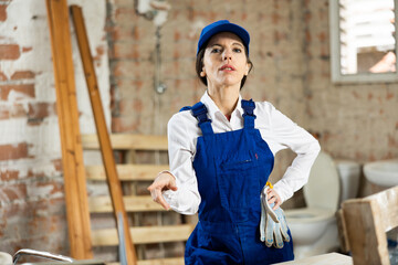 Confident positive female builder wearing blue overalls and cap posing at construction site...