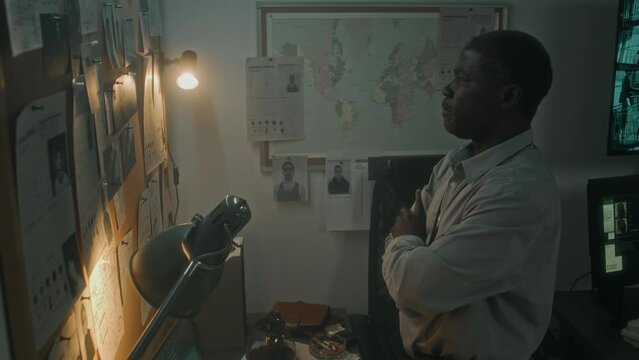 Medium arc shot of African American crime scene investigator standing in office, looking intently at pin board with photos and police bulletins for suspects, analyzing murder and contemplating motive