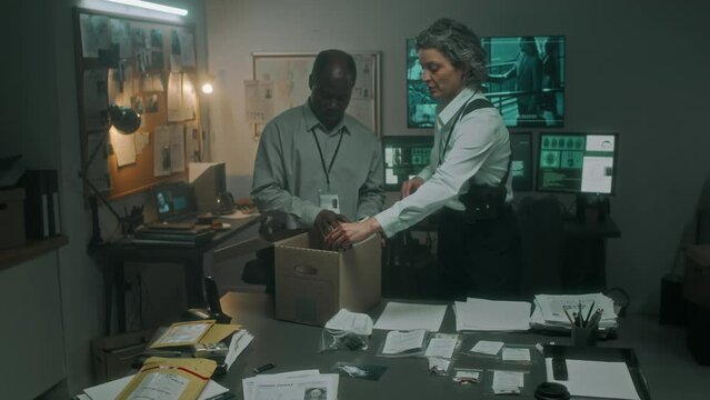 Medium full shot of African American male and Caucasian female special agents reviewing evidence laid out on table, bringing in boxes with case files and searching through investigation documents