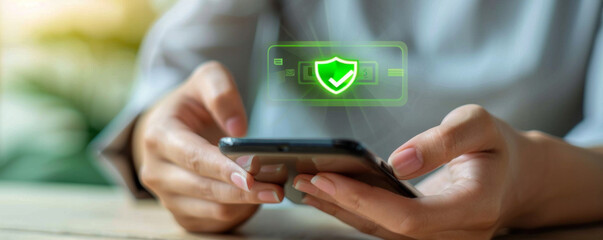 A woman is holding a cell phone and looking at a green shield on the screen. The shield is a symbol of protection and security