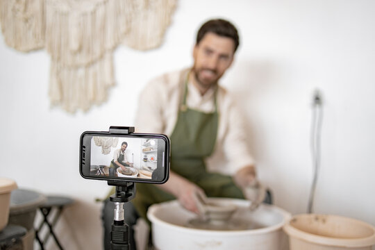 Mature man shaping clay vase using potter's wheel and recording video blog with smartphone. Craftsmanship and creativity showcased. - Powered by Adobe