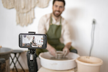 Adult male potter creating pottery on a potter's wheel while recording a video blog using a...
