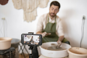 Young adult male potter sculpting vase on potter's wheel while recording video blog using smartphone on tripod.