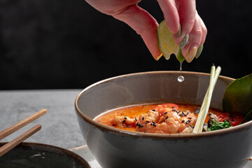 A popular dish of Asian cuisine is Tom Yam soup, cooked with shrimp, lemongrass, galangal and lime. A hand squeezes a drop of lime into a Thai soup with a sour-spicy taste.