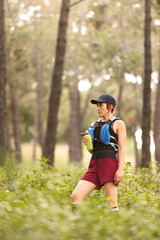 A woman wearing a red jacket and blue vest holding a bottle and a water bottle. She is wearing a black hat