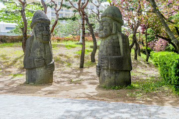 Two large stone grandpa statues (Dolhareubang) in a garden area beside walkway with trees and bushes