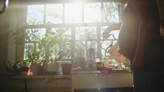 Older man pouring water in kitchen with plants on windowsill. Natural light from windows in the background. Domestic life and morning routine concept. Design for poster, banner, greeting card. Candid