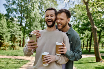 A bearded gay couple shares a laugh while looking at a phone and enjoying coffee in a green park.