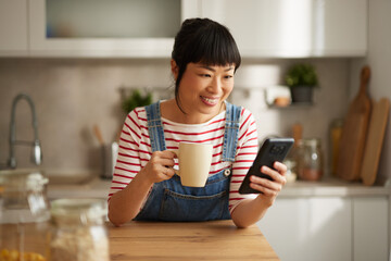 Beautiful Asian woman using mobile phone while drinking coffee in the kitchen