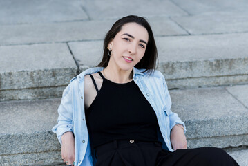 Fototapeta premium Attractive Caucasian Young Woman Brunette Hippie In Casual Clothes Posing With Pleasure While Walking Outdoors On A Warm Spring Day Sitting On The Steps. Stylish girl.