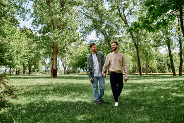 Fototapeta premium A bearded gay couple walks hand-in-hand through a lush green park, enjoying a sunny day together.