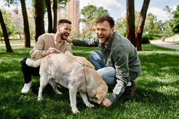 A bearded gay couple laughs while playing with their labrador retriever in a green park on a sunny...