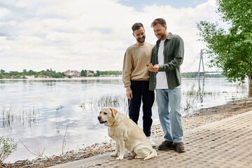 A bearded gay couple enjoys a walk with their labrador dog by a lake on a sunny day.