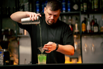 a male bartender pouring a alcoholic drink from the steel shaker to the glass through the sieve on...