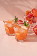 Glasses with summer tropical fruit cocktail drinks with sliced peaches and ice on pastel pink background with bright natural sun light and floral shadows