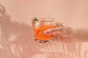 Hand holding glass with iced summer refreshing drink with peach fruits and ice, on pastel peachy pink background with natural sunlight shadows, lifestyle