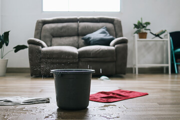 Water dripping from ceiling into plastic bucket on floor in living room. Leaking roof concept.
