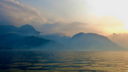 Morning over a tropical island. View from the sea of ​​a hilly tropical island shrouded in fog and the surface of a calm sea in the rays of dawn.