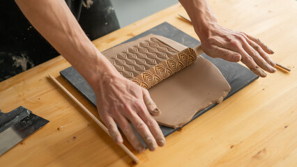 Close-up of a potter's hands rolling out clay using a rolling pin with patterns. 