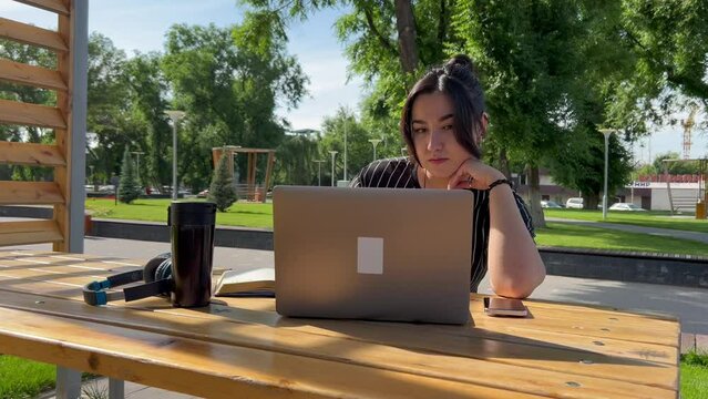 young brunette Asian woman student or freelancer working with laptop in the park outdoors on a sunny summer day