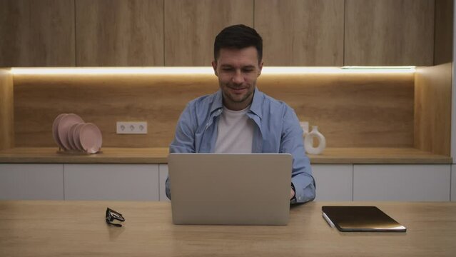 A man, working from home using a laptop and a tablet, smiles as he works as a freelance online educator. He wears glasses and has a professional appearance.
