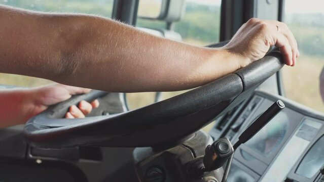 Male hands of trucker holds a big steering wheel while operating a truck through countryside. Lorry driver riding to destination at country road. Concept of logistics and transportation. Close up