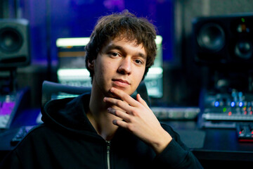 in dark recording studio against the backdrop of musical equipment for sound processing, a young guy sits looking at the camera and gesticulating