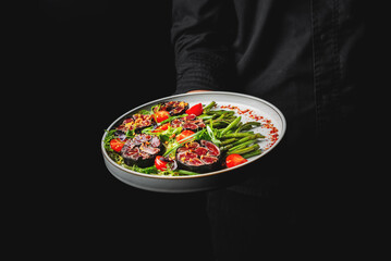 A fresh salad with green beans, tomatoes, and meat on a plate held by a person against a black background