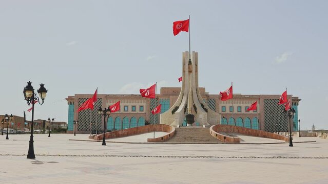 Sun-drenched Kasbah Square, huge paved esplanade in center of Tunis with National Monument surrounded by red Tunisian flags, symbolizing national pride and history, against City Hall building. High